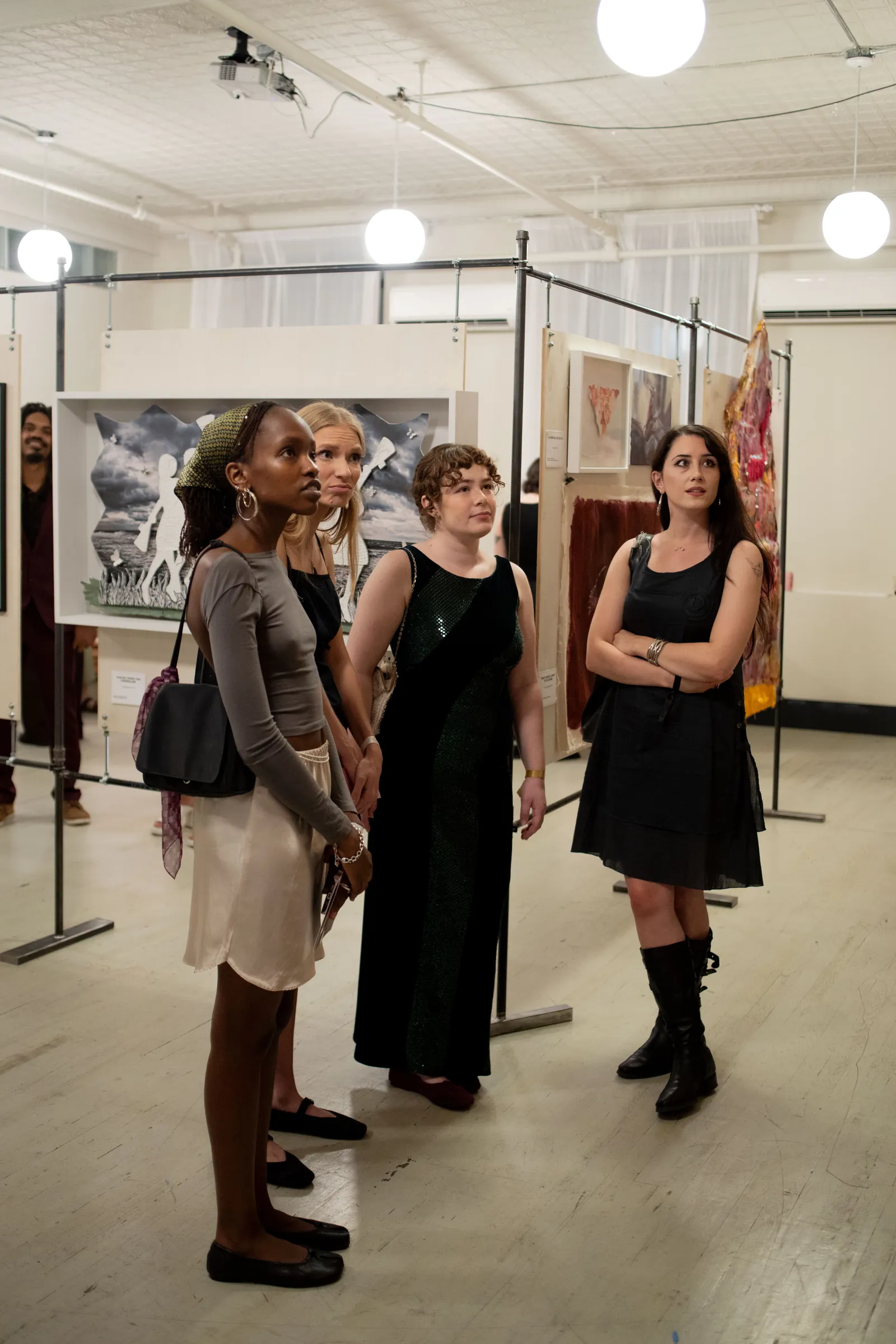 Four visitors stand together looking up at a work just out of frame, beneath globe pendant lights, with a paper-cut artwork and textile pieces on the panels behind them.