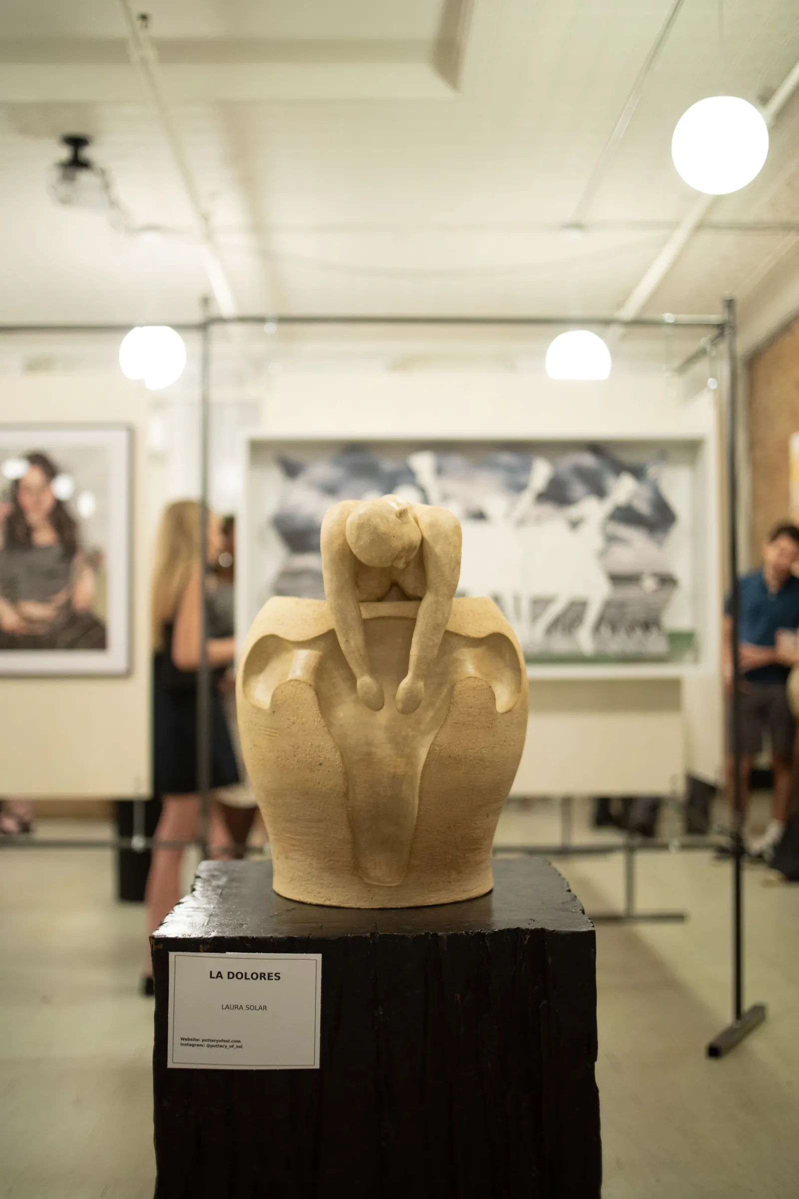 La Dolores, a ceramic sculpture by Laura Solar: a small figure folds over the rim of a vessel carved in the shape of a uterus, set on a dark wooden plinth with the exhibition blurred behind.