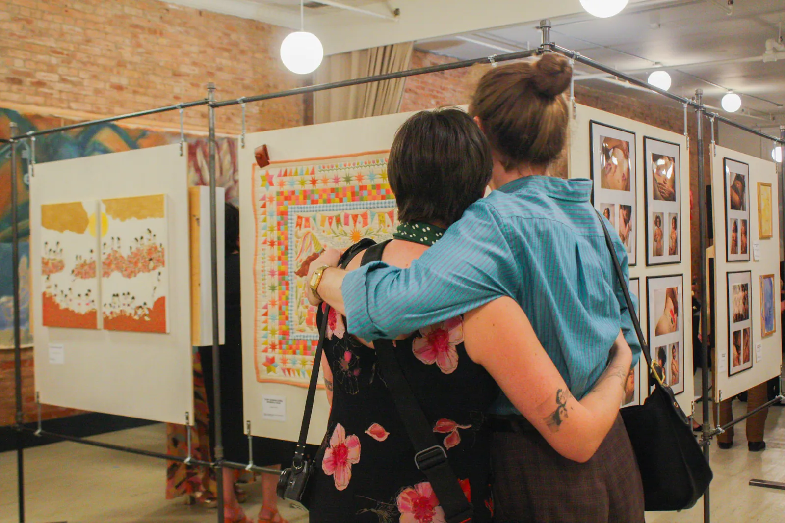 Two visitors, seen from behind, stand arm in arm before a bright star-patterned quilt, framed photographs and a yellow diptych around them.