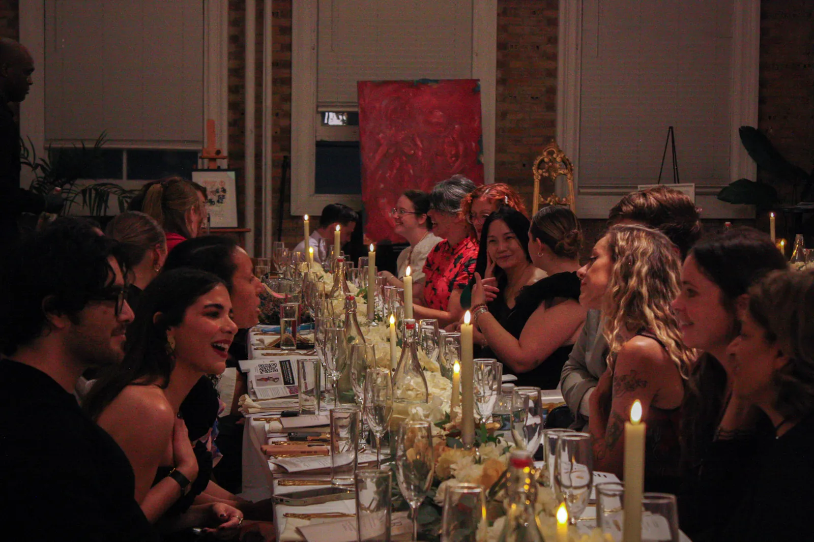 Guests lean toward one another across a table of candles and white flowers, a red painting on an easel behind them.