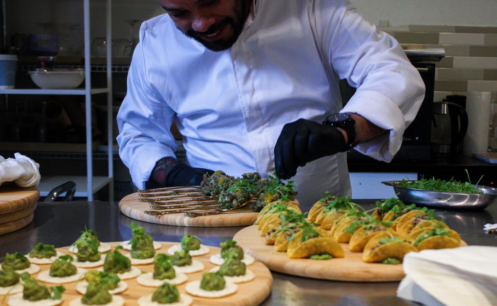 A chef in a white jacket smiles as he garnishes trays of canapés and small tacos with microgreens in the kitchen.