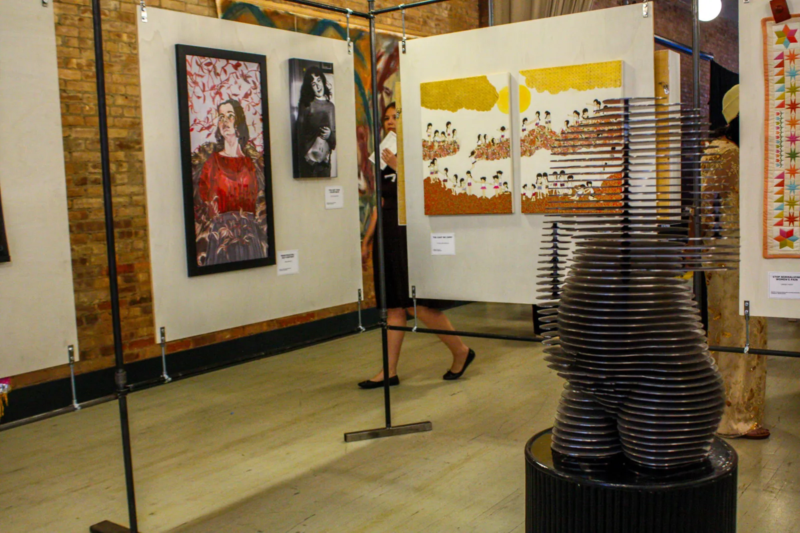 An installation view of the Annex: a stacked-disc sculpture in the foreground, a red figurative painting, a black-and-white photograph and a yellow diptych on the panels behind, a visitor passing through.