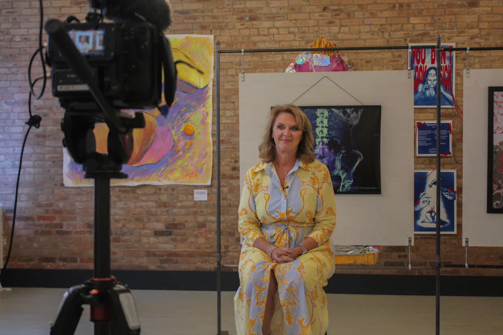 A woman in a yellow and blue patterned dress sits for a filmed interview among the artworks, a camera in the foreground.