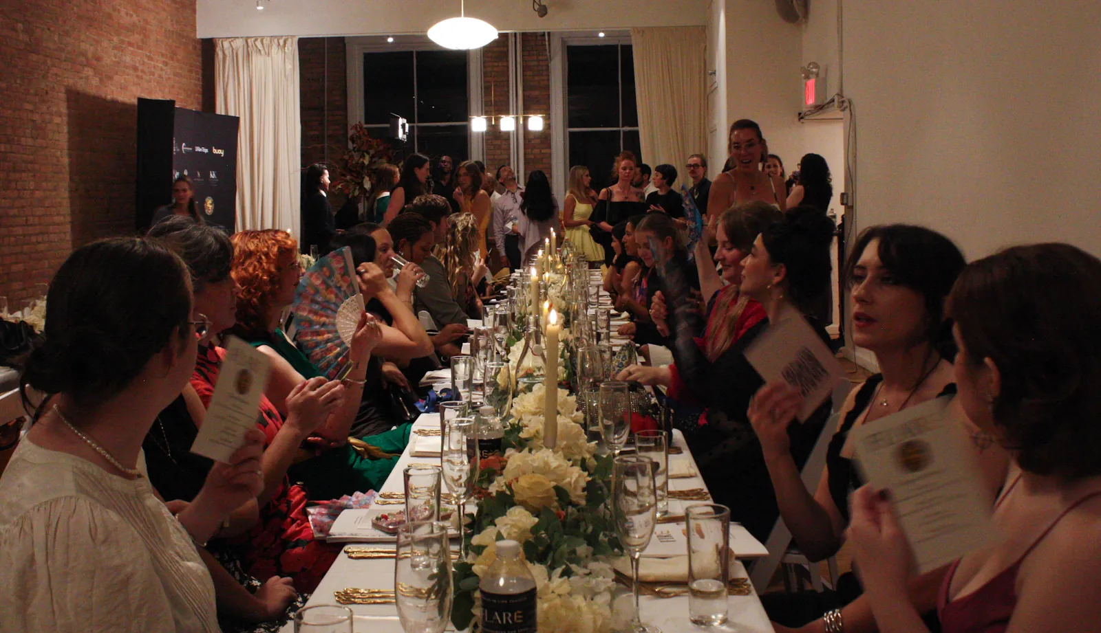 A long candlelit dinner table fills the room, guests seated on both sides and more standing beyond, some fanning themselves with menus.