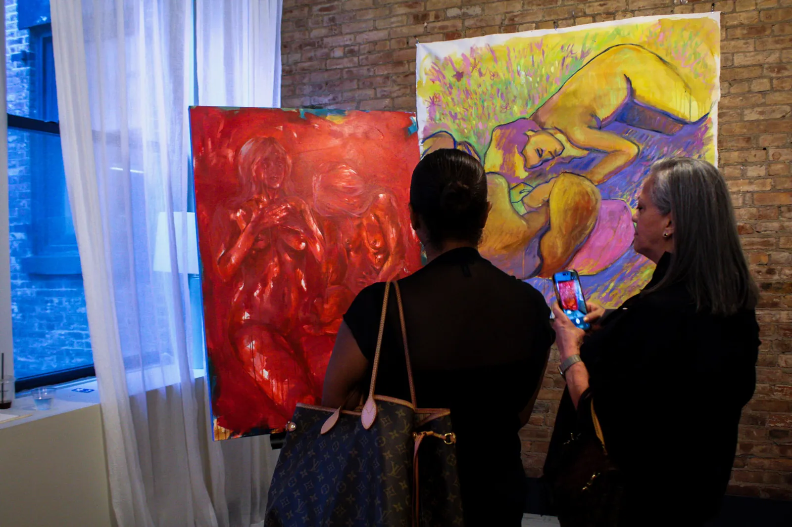 Two visitors seen from behind stand before a red figurative painting and a large yellow and violet canvas; one photographs the work on her phone.