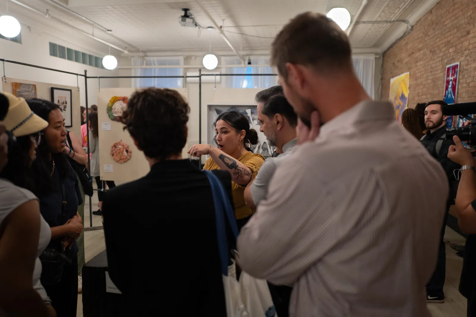 Laura Solar, in a mustard top, points toward her ceramic wall pieces as a close circle of visitors listens and a videographer films.