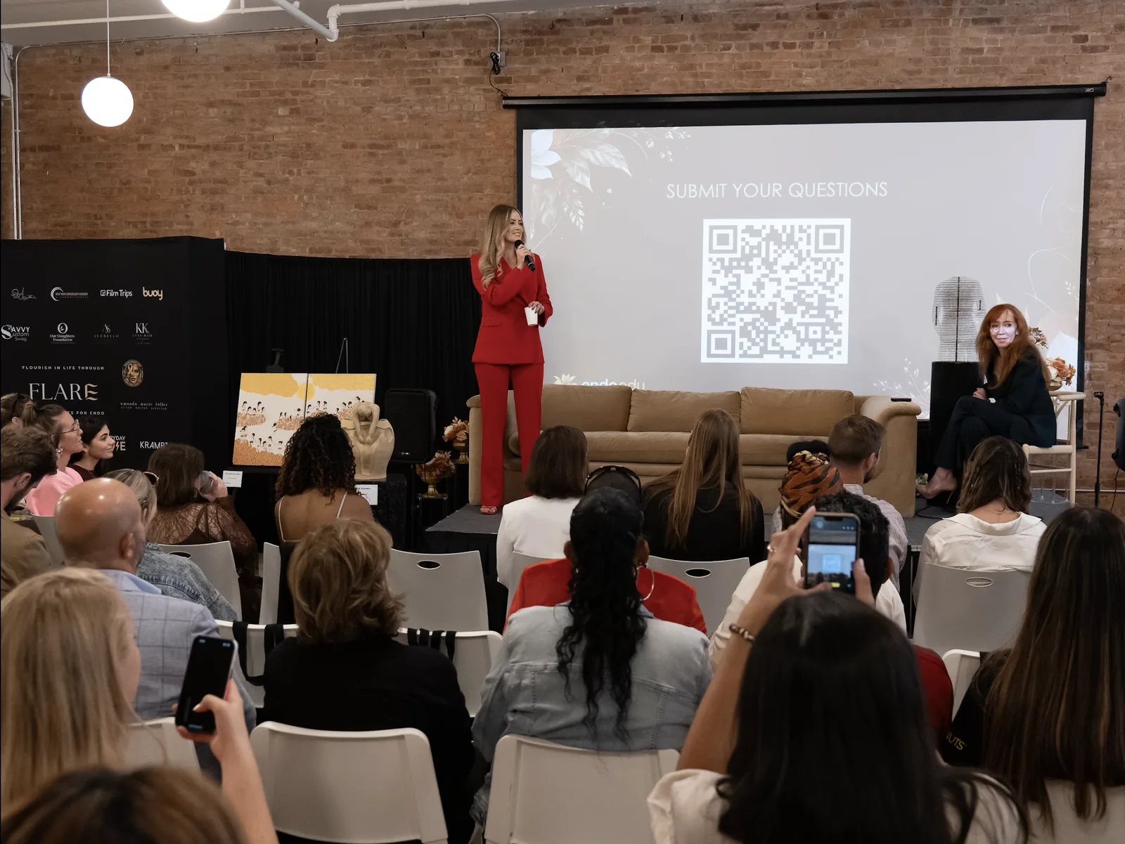 Savannah Regensburger, in a red suit, speaks to a full room during the joint keynote while Dr. Chandra Spring-Robinson listens from a stool beside the stage; a screen behind them reads Submit your questions.