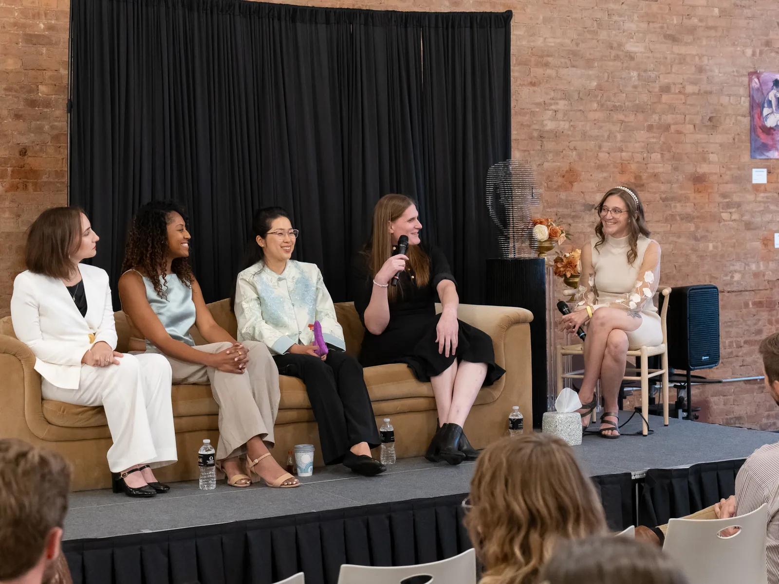 Five women on a low stage share a panel, four on a velvet sofa and the moderator on a chair, smiling as one of them speaks into a microphone.