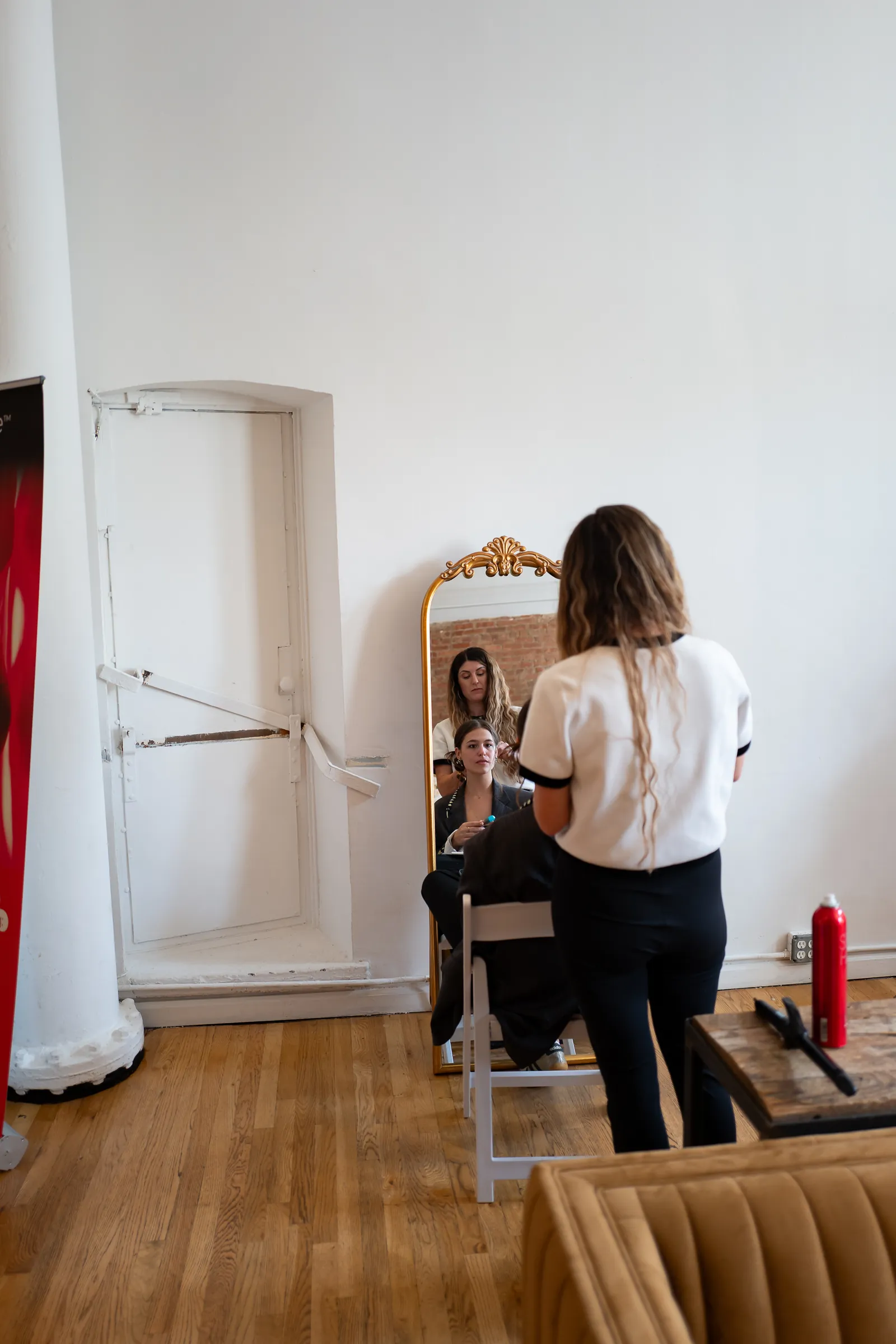 In the Self-Care Lounge, a stylist works on a seated guest’s hair, both reflected in a tall gold-framed mirror.