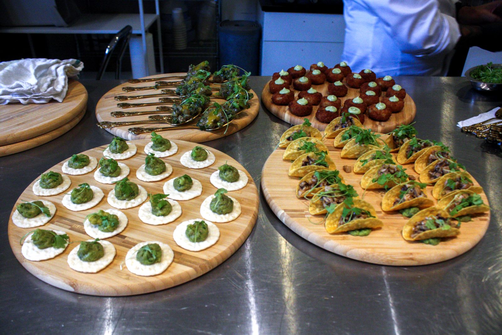 Four wooden boards of passed hors d’oeuvres on a steel counter: avocado bites, gold spoons, croquettes and mini tacos