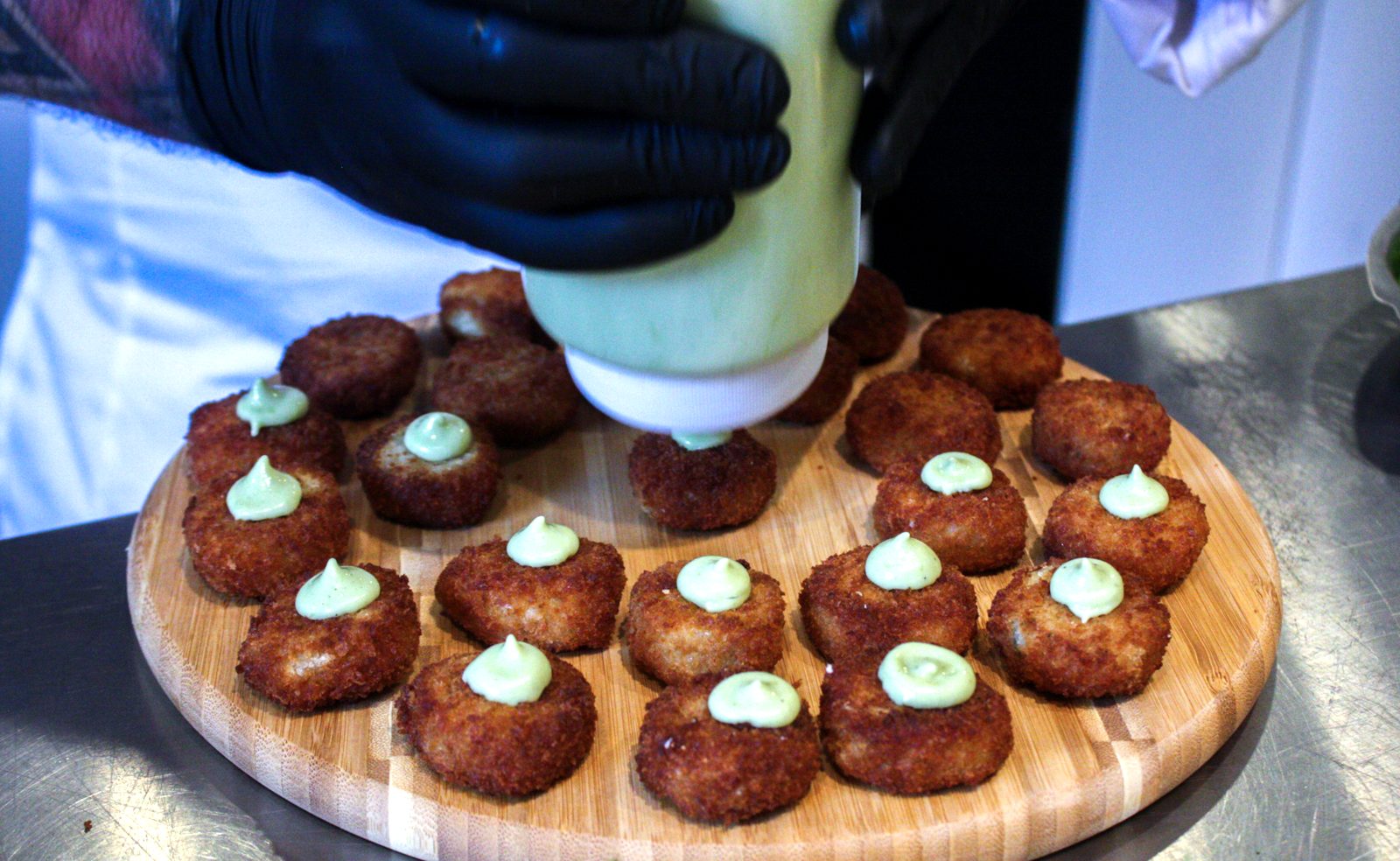 A gloved hand pipes pale green sauce onto golden croquettes