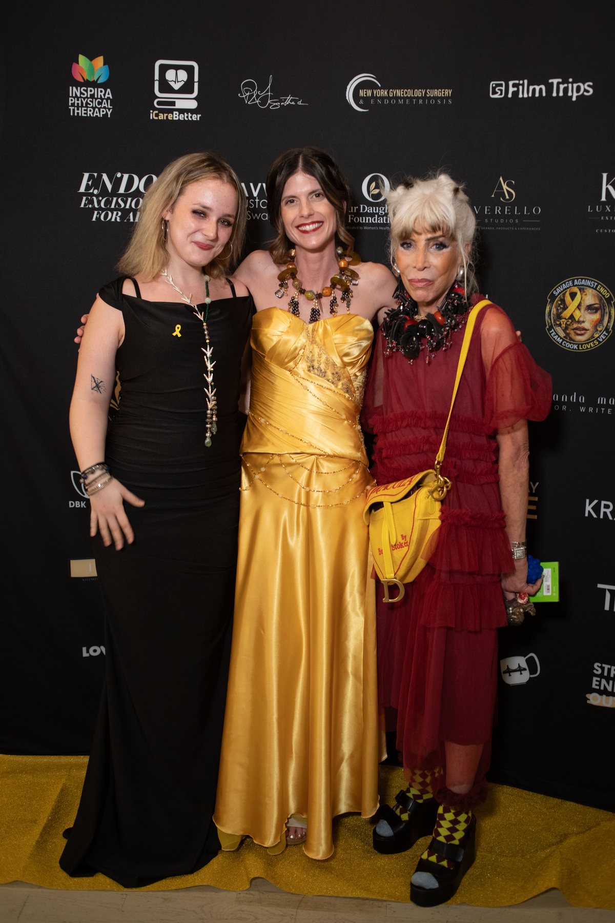 Alexandra Totillo, Priscilla Smith and Sara Gay stand together on the gold carpet in front of the sponsor wall; Priscilla wears the gold dress and a large beaded necklace.