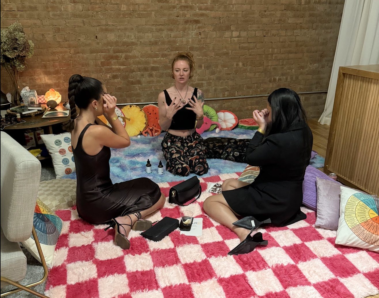 Elizabeth Snider leads two guests through a somatic practice, seated on cushions and a pink-and-white rug in the Self-Care Lounge