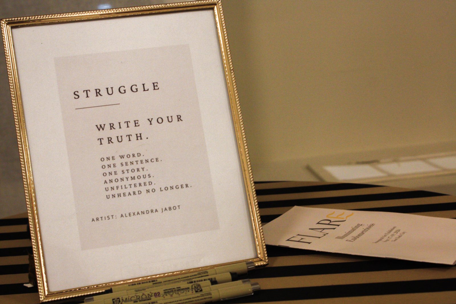 Framed sign: Struggle. Write your truth. One word. One sentence. One story. Anonymous. Unfiltered. Unheard no longer. Artist: Alexandra Jabot