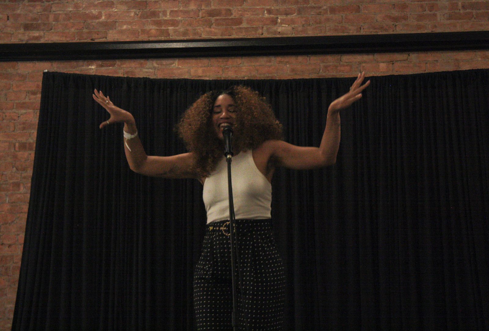 Ariel Bridges on stage, arms raised and eyes closed, singing in front of a black curtain and brick wall