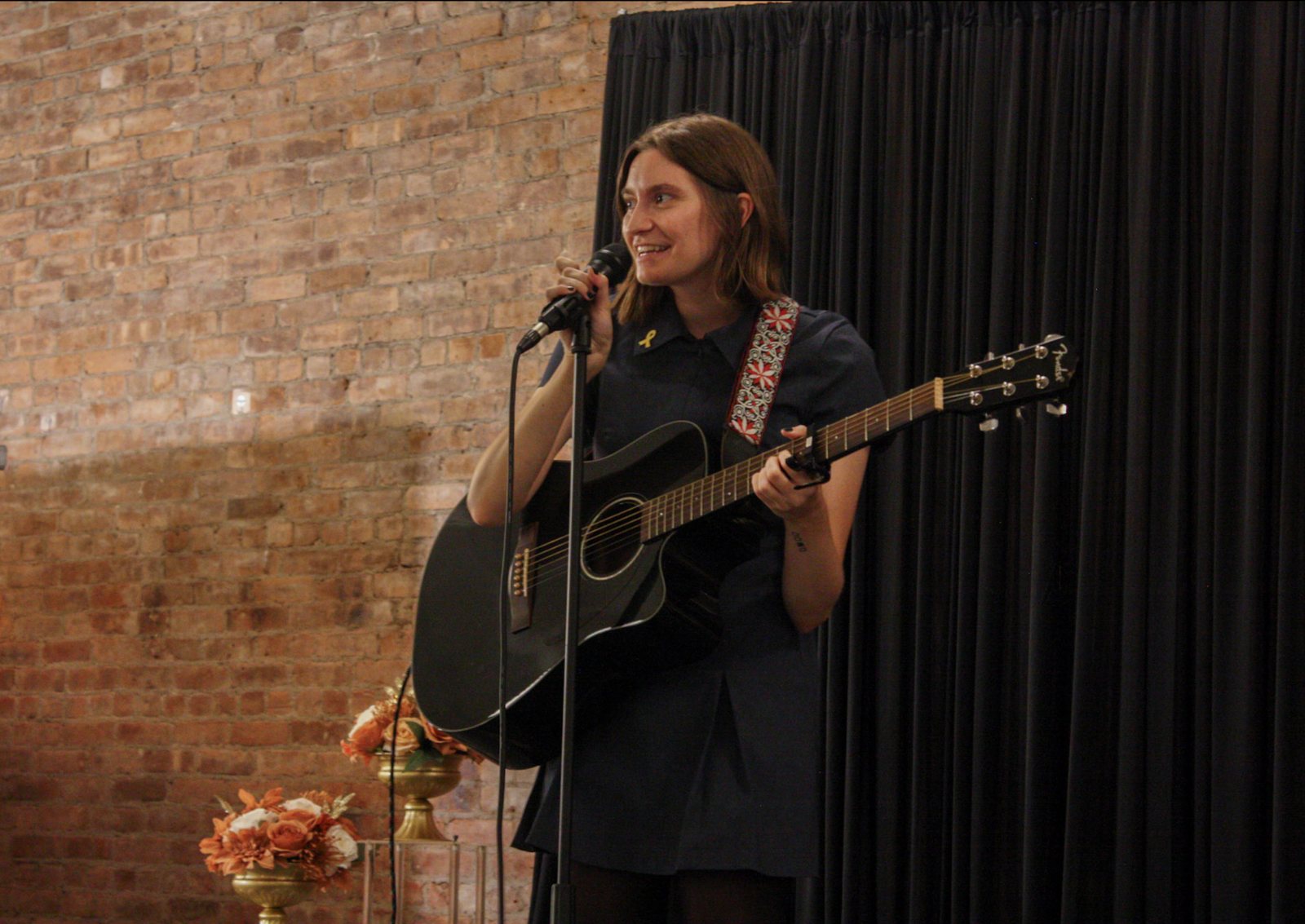 Carrie Wallace with a black acoustic guitar, smiling as she speaks into the microphone