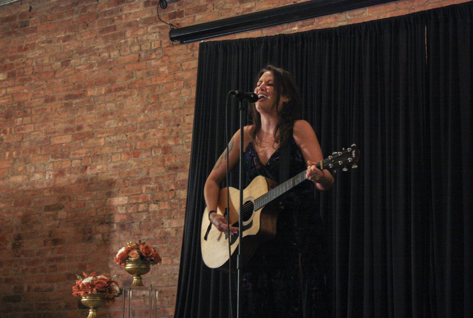 Debra Stark singing and playing an acoustic guitar in front of a black curtain and brick wall